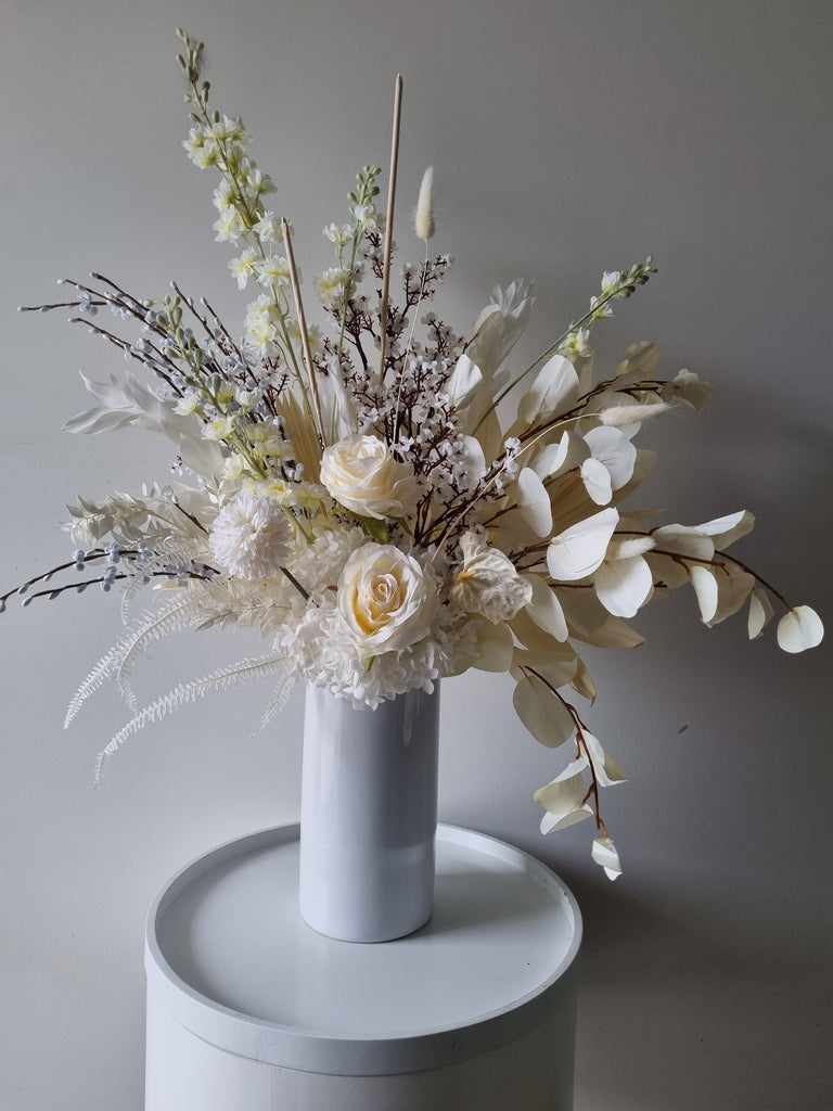 Elegance floral arrangement containing white and cream artificial and dried flowers with foliage, presented in a tall white vase.