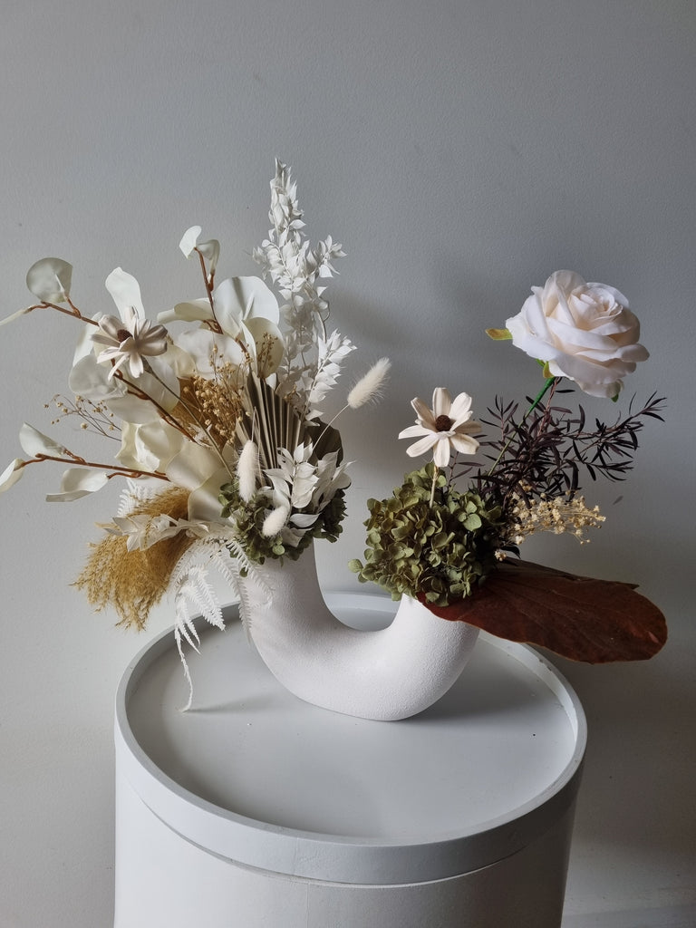 Forest floral arrangement containing soft pink, earthy and neutral coloured flowers and foliage, presented in a white U-shaped vase with two openings.