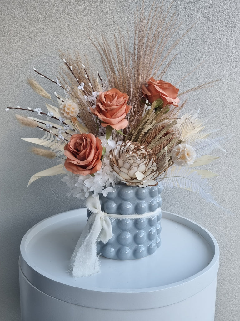 Bubbles floral arrangement containing white, soft orange and neutral artificial and dried flowers as well as foliage, presented in a bubbly baby blue vase.