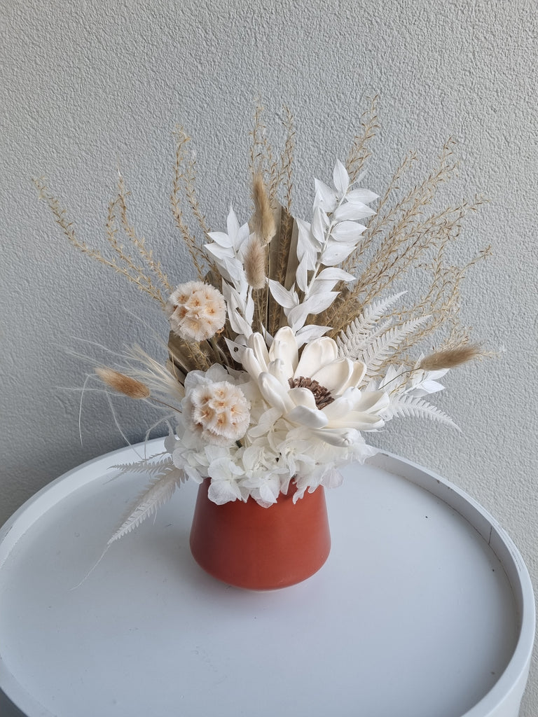 Terracotta floral arrangement made with cream and white artificial flowers with foliage, presented in a terracotta orange vase.