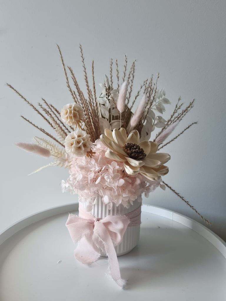 Petite floral arrangement made with cream and soft pink artificial and dried flowers with foliage, presented in a small white vase with matching ribbon.